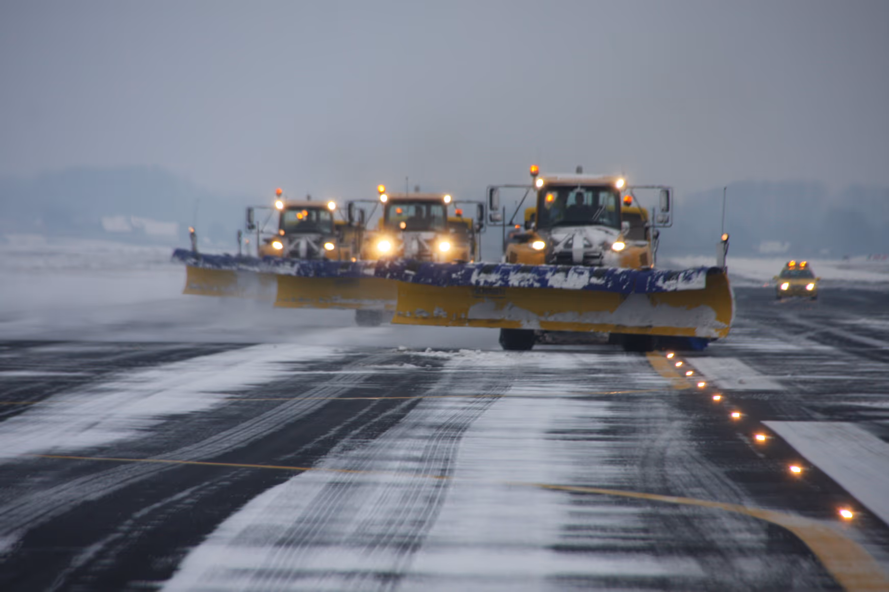 Four airport snowplows with headlights on clear airfield pavement in blowing snow, leaving streaks of slush beside a line of lit in-pavement lights.