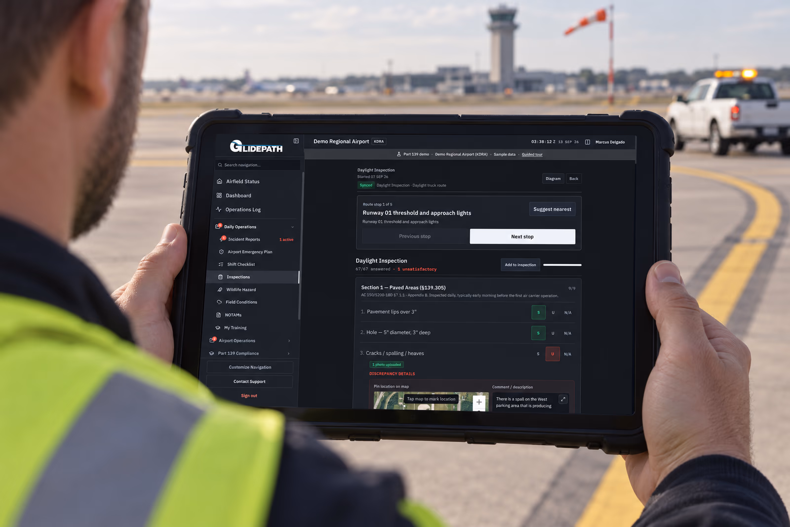 Over the shoulder of an airport operations worker in a hi-vis vest on the apron, holding a rugged tablet that shows a Glidepath daylight self-inspection: the route stop, the checklist with a paved-areas item marked unsatisfactory, a photo uploaded, and a map pin for the discrepancy, with an operations truck and the control tower behind.