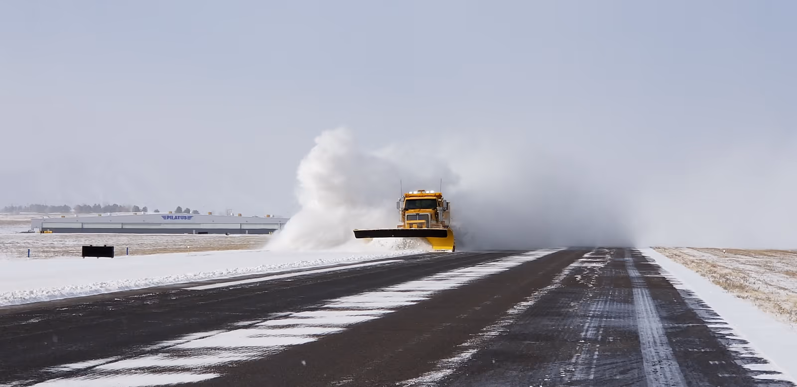 A snowplow throws a plume of snow down a runway, with a cleared dark strip beside lanes still streaked with snow.