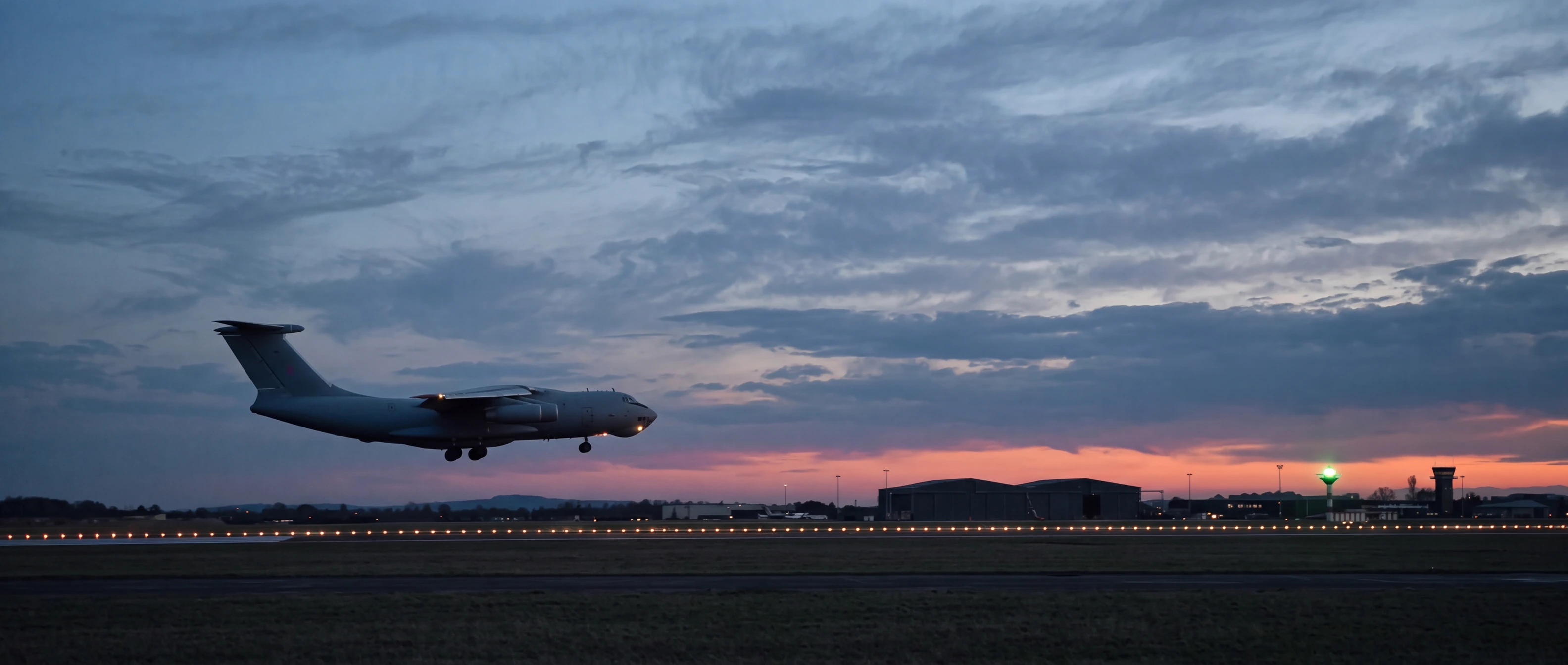 A grey military transport in side profile on short final at blue hour, descending left to right toward the glowing runway edge lights under dramatic dusk clouds