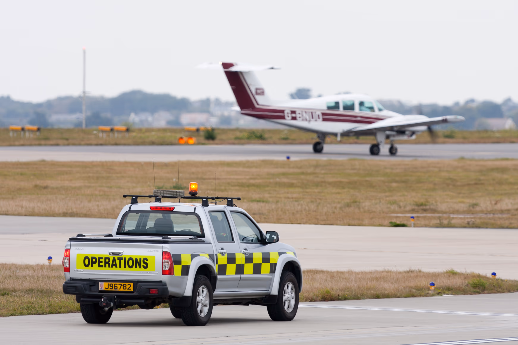 An airport operations pickup with an amber beacon on its roof waits on a taxiway beside the runway, as a light twin-engine aircraft taxis past.